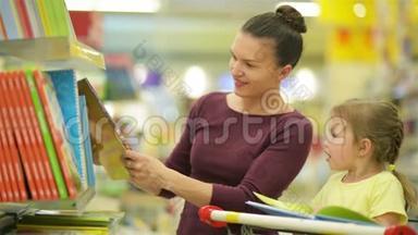 年轻的母亲和女儿在超市挑选书籍。 漂亮的女儿坐在超市的手推车里